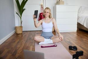 Portrait of beautiful lifestyle blogger, recording herself working out at home, sitting on rubber mat doing yoga in her room, taking selfie on smartphone, using digital camera photo