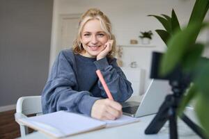 linda sonriente niña se sienta en un habitación, escribe abajo notas, haciendo tarea, registros de sí misma en digital cámara, crea contenido para vlog, estilo de vida blogger haciendo diario rutina episodio foto