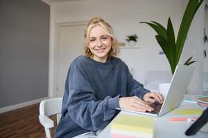 retrato de sonriente joven mujer, Universidad estudiante se sienta en su habitación, lo hace tarea, estudios remotamente desde hogar, usos ordenador portátil para Lanza libre trabajo foto