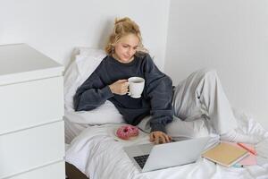 retrato de joven hermosa rubio mujer, sensación perezoso a hogar, acostado en cama con taza de té, rosquilla y computadora portátil, estudiando remotamente o acecho película en línea foto