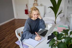 Lifestyle image of young woman writing down something in notebook, making notes, studying online, doing course in internet, listening to interesting information, using laptop photo