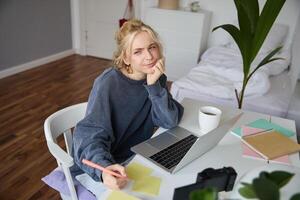 Portrait of young thoughtful girl, studying, making notes, writing down something in notebook, doing homework in her room, sitting in front of laptop, frowning while thinking photo