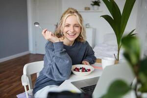 imagen de riendo mujer sentado en frente de ordenador portátil en su habitación, comiendo desayuno, participación cuchara y cuenco en mano, acecho s en línea mientras teniendo un bocadillo foto