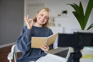 Portrait of cute, stylish young woman, shows okay sign, holds notebook, reads aloud her notes, records vlog, looks at digital camera and makes ok gesture photo