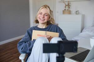 Portrait of smiling, charismatic young woman, content creator, records on digital camera, laughing and looking happy, using journal, writing in notebook photo