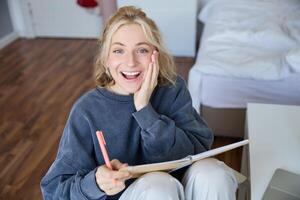 Portrait of happy, young blond woman, sitting in bedroom with notebook and pen, laughing and smiling, writing in journal, doing homework and smiling photo