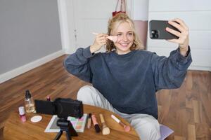 Image of stylish young woman, social media influencer, taking pictures on mobile phone, doing makeup tutorial for followers online, recording vlog in her bedroom, showing brush photo