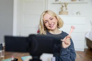 Close up portrait of happy young beauty blogger, records lifestyle vlog in her room, using camera with stabiliser, shows makeup brush and cosmetics photo