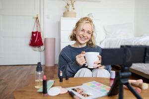 retrato de sonriente joven mujer, niña registros en cámara, sostiene taza de té, hablando, haciendo estilo de vida Blog, sentado en habitación y creando contenido foto