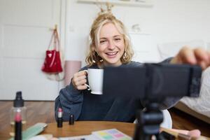 retrato de linda sonriente mujer, rubio niña bebidas té y registros un casual, estilo de vida Blog, vlogger se sienta en un habitación con cámara y estabilizador, sostiene taza de café y negociaciones foto