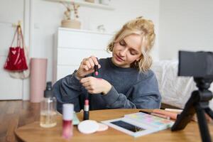 Woman beauty blogger, records of herself sitting in a room and rating makeup products, puts on make up, holds lipstick and cosmetic brush in hand, using professional camera for content creation photo