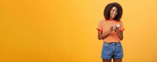 Stylish carefree girl texting friend come over standing pleased over orange wall in stylish denim shorts typing message or scrolling news in internet via smartphone gazing at device screen with smile photo