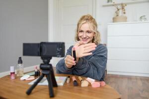 Portrait of young blond woman, teenage girl records for her social media account, shows makeup on camera, recommends lipstick to online followers, creates content in her room photo