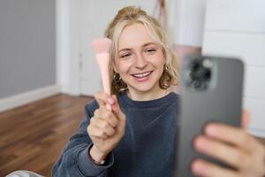 Image of stylish young woman, social media influencer, taking pictures on mobile phone, doing makeup tutorial for followers online, recording vlog in her bedroom, showing brush photo