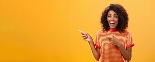 Portrait of amazed excited charismatic dark-skinned young pretty girl with afro hairstyle in trendy striped t-shirt pointing left delighted and fascinated posing against orange background photo
