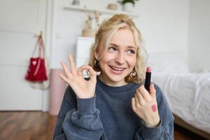 Young female blogger, content creator showing lipstick and okay hand sign, recommending beauty product for her audience on social media, recording vlog in room photo