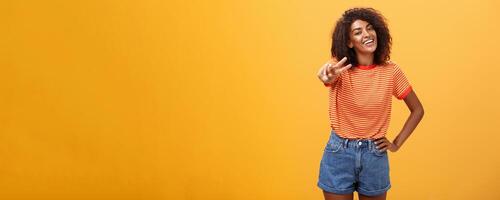 Friendly carefree and happy good-looking tall african american female model with curly hairstyle holding hand on hip tilting head and smiling joyfully showing peace gesture over orange wall photo