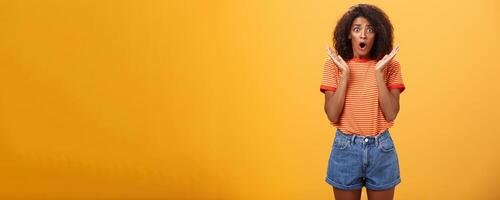 Oh no what happened. Worried african american sister with curly hair in trendy striped t-shirt and denim shorts gasping dropping jaw, looking concerned expressing empathy and shock over orange wall photo