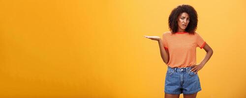 So what. Moody unimpressed stylish dark-skinned girlfriend with curly hairstyle holding hand on waist frowning and raising palm in your point gesture being confused and questioned over orange wall photo