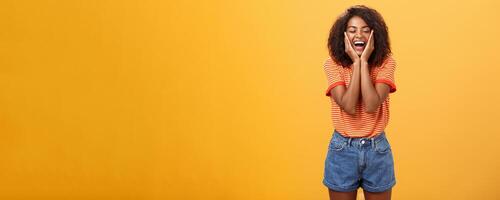 Woman happy and delighted with great result after curing acne with skincare products touching cheeks smiling happily with closed eyes feeling pleased and amused over orange background photo