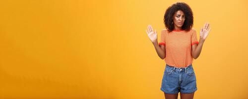 Silly insecure and sad dark-skinned female model in trendy striped t-shirt and shorts raising arms in surrender frowning being uninvolved and unaware standing clueless over orange background photo