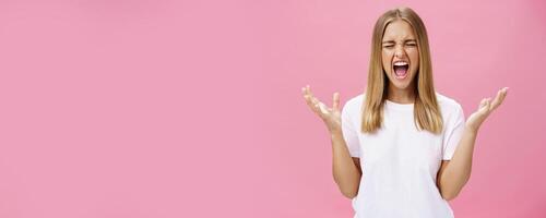 Woman feeling fed up having mental breakdown during deadline yelling with closed eyes, raising hands aside in pissed gesture feeling distressed and pressured losing temper over pink wall photo