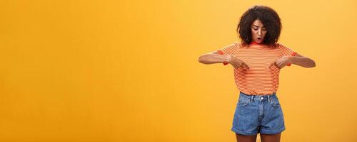 Portrait of worried questioned and surprised african american female with curly hairstyle looking and pointing at stomach or belly feeling discomfort and problems with health over orange background photo