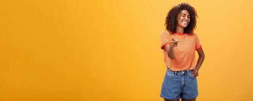 Girl pointing at funny and hilarious friend approving good joke. Amused and entertained attractive dark-skinned female with afro hairstyle laughing with closed eyes indicating at camera joyfully photo