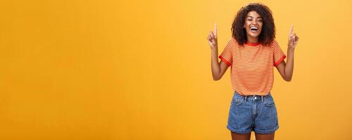Woman feeling amused and entertained. Portrait of happy carefree stylish African-American girl with afro hairstyle laughing out loud joyfully pointing up with raised arms over orange wall photo