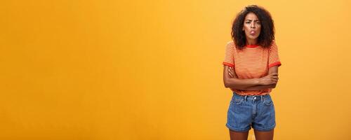 Immature girl showing bad side of character. Portrait of childish offended or displeased young African-American woman with curly hair showing tongue crossing arms on chest over orange background photo