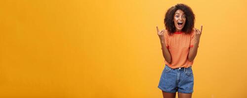 Stylish african american woman being delighted and excited before cool concert showing rock n roll gesture with both hands screaming from satisfaction and happiness cheering over orange background photo