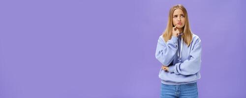 Silly concerned girlfriend trying think up how solve problem standing thoughtful with troubled expression touching jaw pouting, frowning staring right, thinking making up decision over purple wall photo