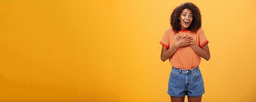 Cute delighted african american woman being grateful and thankful for help standing pleased over orange wall holding palms on breast thanking friend for rescuing grinning joyfully at camera photo