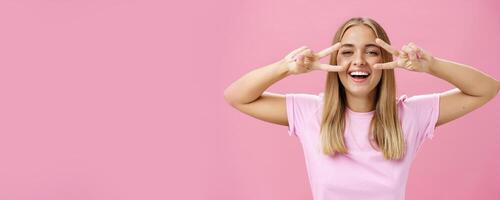 Joyful friendly and peaceful common european woman with blond hair in casual t-shirt showing peace or disco signs over eyes smiling cheerfully at camera having fun against pink background photo