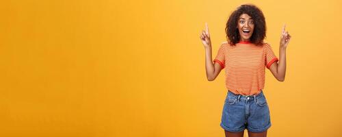 Charmed and impressed african american stylish woman seeing incredible and awesome item on sale raising hands and pointing up with broad grin being excited of new purchase over orange wall photo