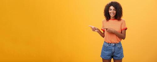 Portrait of charming friendly-looking african-american female coworker in casual outfit pointing left and smiling broadly showing perfect copy space for customers against orange background photo