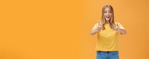 Woman supports with raised thumbs up and amused cheerful smile showing positive attitude expressing like on concept or idea giving approval posing happy and delighted against orange background photo