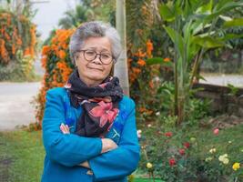 Portrait of an elderly Asian woman wearing a sweater, scarf, glasses, and looking at the camera while standing in the garden. Space for text. Concept of aged people, winter season and healthcare photo