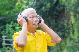 Senior man wearing wireless headphones for listening to a favorite song, and looking up while standing in a garden. Concept of aged people and relaxation photo
