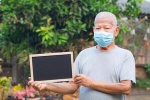 Senior man wearing a mask and holding a blackboard while standing in a garden. Mask for protecting virus, coronavirus, covid-19, pollen grains, and more. Concept of aged people and healthcare photo