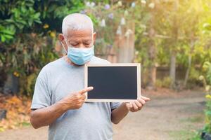 mayor hombre vistiendo un mascarilla, participación y punto a un pizarra mientras en pie en un jardín. máscara para proteger virus, coronavirus, COVID-19, y más. concepto de Envejecido personas y cuidado de la salud foto