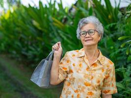 An elderly Asian woman wearing glasses smiling and holding a shopping bag while standing in the garden. Space for text. Concept of aged people and shopping photo