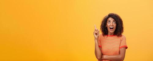 Waist-up shot of interested curious good-looking dark-skinned female in striped t-shirt talking asking question about curious star looking and pointing up with joy over orange background photo
