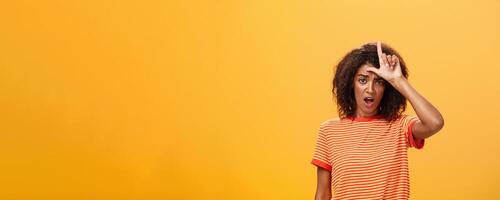 Girl thinks she loser. Portrait of gloomy bothered and displeased african american woman with afro hairstyle showing l word over forehead complaining feeling gloomy and unhappy over orange background photo