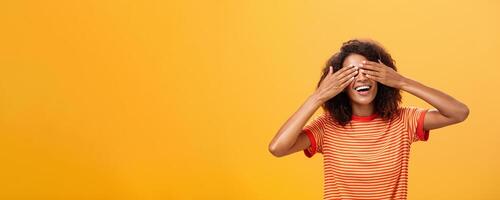 Closing my eyes and counting ten. Portrait of charming dreamy and happy funny african american curly-haired female in striped trendy t-shirt covering sight with palm and smiling waiting surprise photo