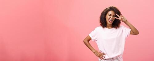 Not afraid express myself. Joyful charismatic african american woman in t-shirt with afro haircut showing tongue playfully and daring making peace sign over eye and winking posing over pink background photo