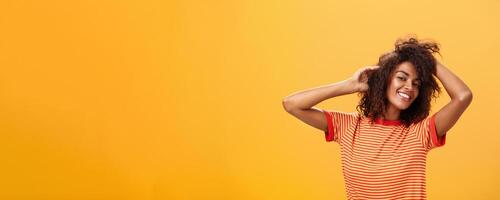 Waist-up shot of charming flirty feminine dark-skinned female in playful mood dancing playing with curly hair and smiling with delight and joy posing over orange background happy and carefree photo