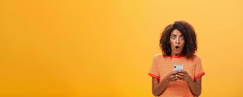 Portrait of shocked and concerned stunned dark-skinned woman with afro hairstyle popping eyes from concern and panic opening mouth holding smartphone revealing terrible information photo