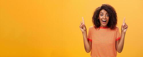 Amazed happy enthusiastic cute african american woman with curly hairstyle in striped t-shirt raising index fingers pointing up gazing mesmerized and delighted at camera over orange wall photo