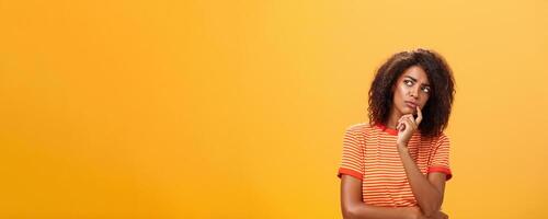 Woman considering to take part in event. Smart focused and creative good-looking african girl with curly hair holding finger on lip gazing concerned left, thinking making decision over orange wall photo